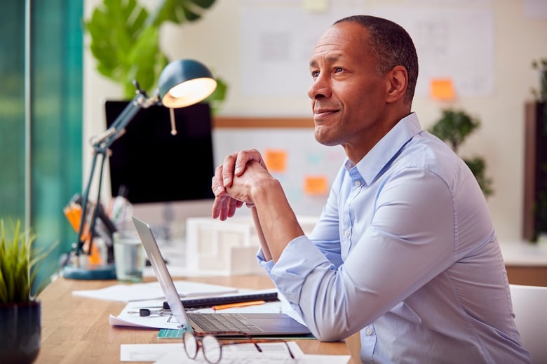 A man in a light blue shirt is sitting at a desk, looking thoughtfully to the side with a slight smile. His hands are clasped together in front of him. The workspace features a laptop, various papers, a pair of glasses, and a desk lamp, with plants and colorful sticky notes in the background, creating a vibrant and inspiring atmosphere.