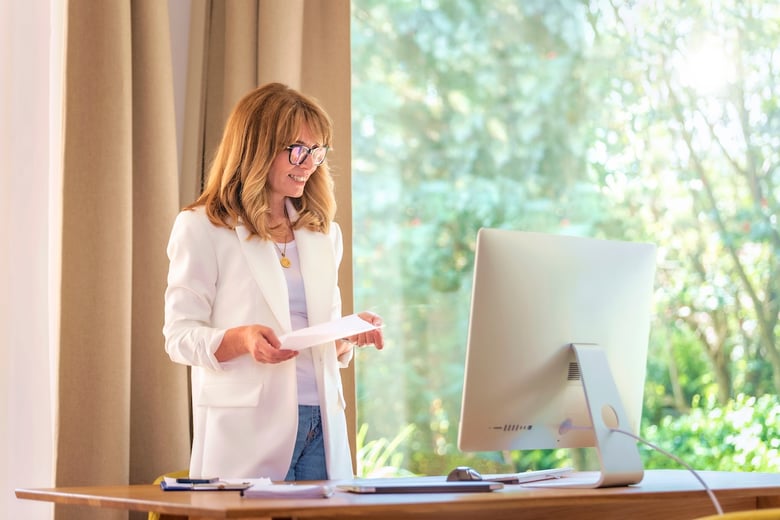 A woman with long, wavy hair is standing at a desk, smiling as she holds a piece of paper in her hands. She is wearing a white blazer over a light top and glasses. In front of her is a computer monitor, and the background features large windows with a view of greenery, creating a bright and cheerful workspace.