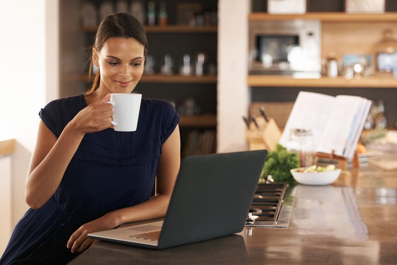 A woman in a navy blue dress is sitting at a kitchen counter, smiling as she holds a white mug. In front of her is a laptop, and in the background, there are kitchen utensils, a bowl of salad, and an open cookbook on a stand. The setting is bright and modern, creating a warm, inviting atmosphere.